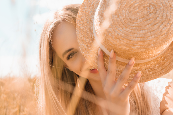 selective focus of blonde woman obscuring face with straw hat while looking at camera  - Fotó, kép