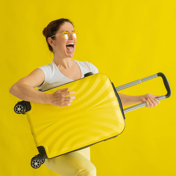Beautiful caucasian woman fooling around with a suitcase on a yellow background. A charming girl imitates playing the guitar. The joy of the journey ahead. - Foto, afbeelding