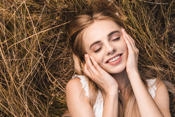 top view of blonde woman touching face while lying on green grass with closed eyes - Foto, Imagem