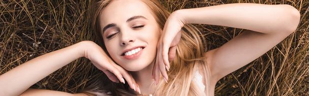 panoramisch concept van sensuele blonde vrouw aanraken gezicht terwijl liggend op gras met gesloten ogen, bovenaanzicht - Foto, afbeelding
