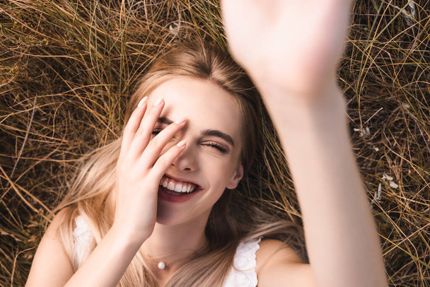 selective focus of excited blonde woman touching face and looking at camera while lying on grass with raised hand, top view - Фото, зображення