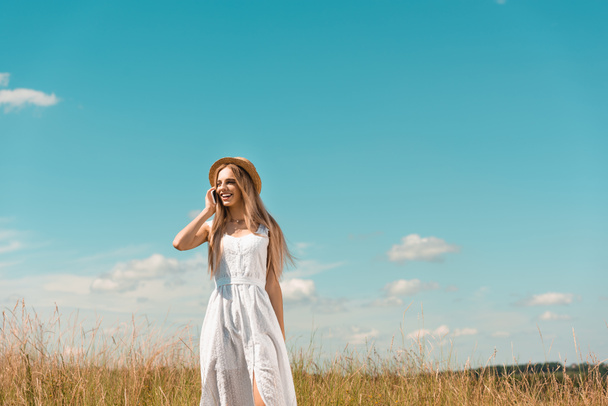stijlvolle blonde vrouw in witte jurk en strohoed praten op smartphone in weide tegen blauwe lucht - Foto, afbeelding
