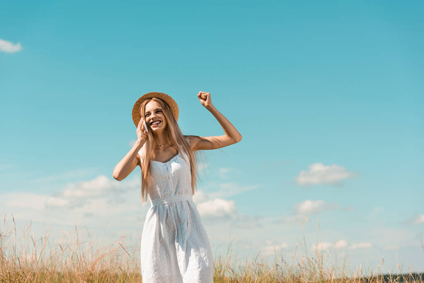 excited blonde woman in white dress and straw hat showing winner gesture while talking on smartphone against blue sky - Fotó, kép
