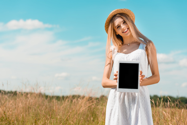 Blondine mit Strohhut und weißem Kleid zeigt im Stehen ein digitales Tablet mit leerem Bildschirm  - Foto, Bild