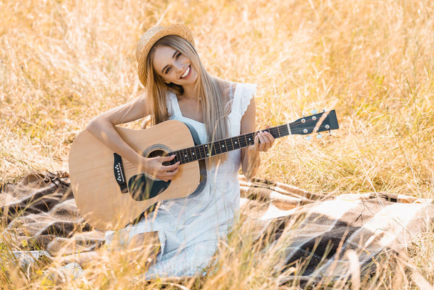 selective focus of young woman in white dress and straw hat looking at camera while playing acoustic guitar on blanket in field - Photo, Image