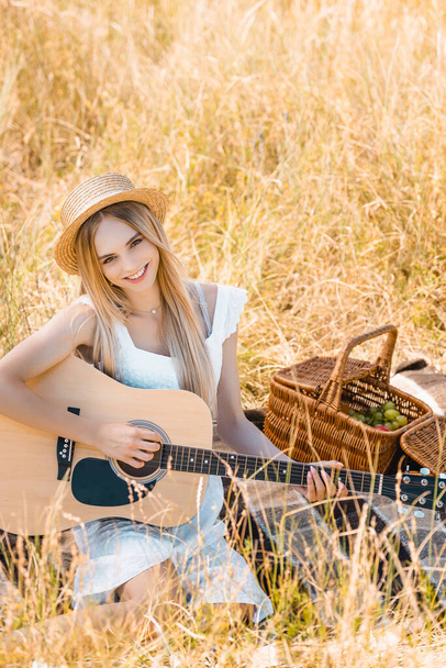 selective focus of blonde woman in straw hat looking at camera while playing acoustic guitar on blanket in meadow, high angle view - Photo, Image