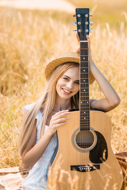 επιλεκτική εστίαση της νεαρής ξανθιάς γυναίκας κοιτάζοντας κάμερα, ενώ κάθεται με ακουστική κιθάρα σε χορτώδες πεδίο  - Φωτογραφία, εικόνα