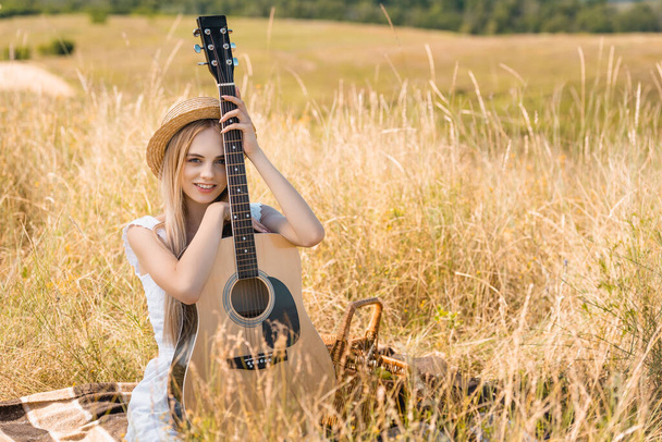 selective focus of blonde woman in summer outfit and straw hat sitting on blanket with acoustic guitar and looking at camera  - Photo, Image