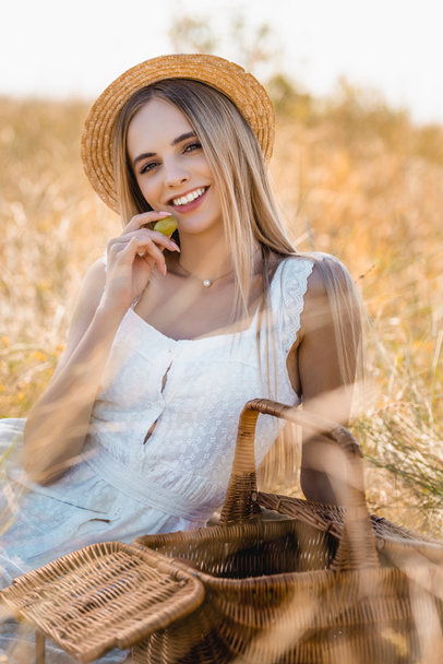 selective focus of blonde woman in white dress and straw hat holding ripe grape while looking at camera in field - Foto, Bild