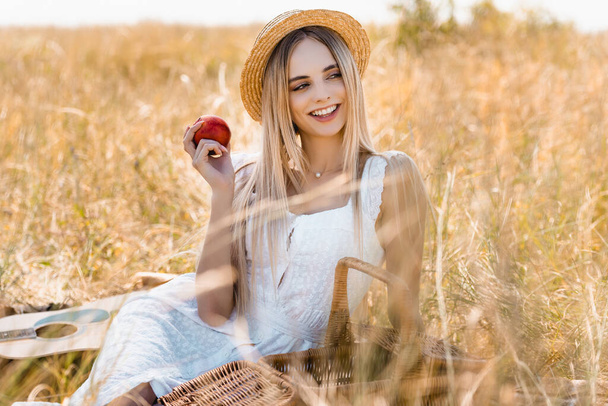selective focus of woman in white dress and straw hat holding ripe apple while sitting in field - Photo, Image