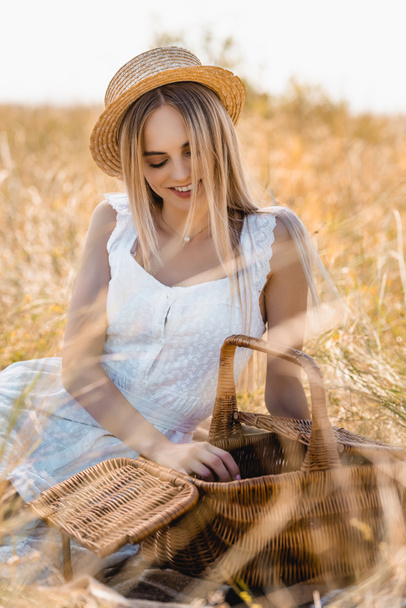 mujer rubia en vestido blanco y sombrero de paja sentado cerca de la canasta de mimbre en el campo - Foto, imagen