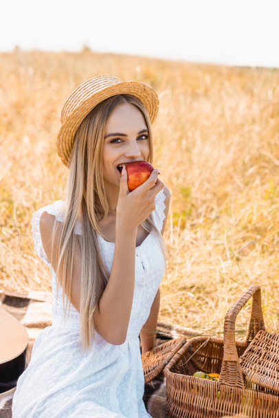 young blonde woman in straw hat eating ripe apple and looking at camera while sitting near wicker basket in field - Foto, Imagen