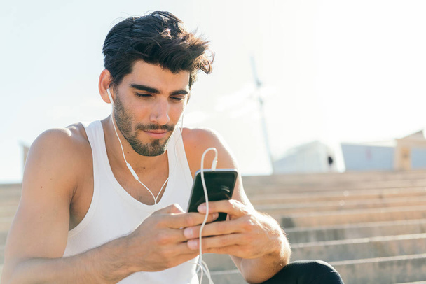 handsome sporty man with earphones selects music on his phone to listen during his workout, urban sport concept and healthy lifestyle, copy space for text - Photo, Image