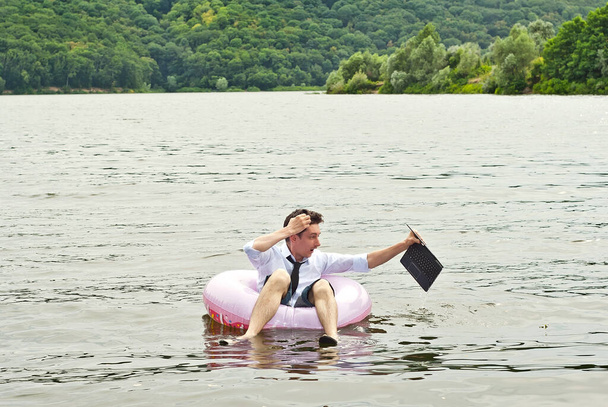 Un hombre con una camisa en un círculo inflable rosa. Un tipo con un portátil nada en el río. El concepto de trabajo durante las vacaciones, outsourcing y freelance. El hombre ahogó la computadora. Infortunio.. - Foto, imagen