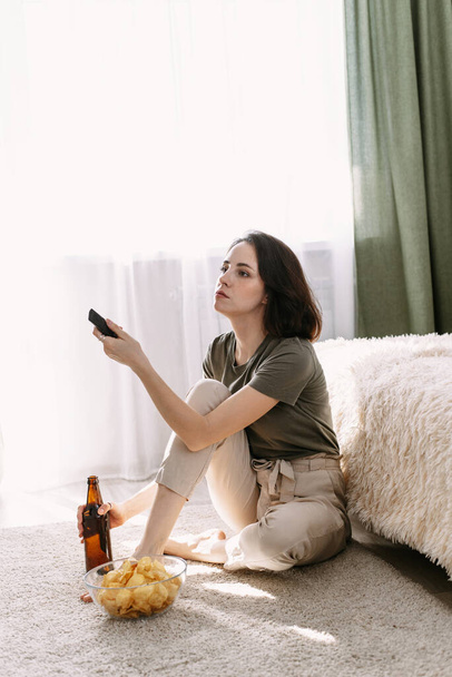 Młoda kobieta pije piwo i je chipsy w domu. Relaks w domu oglądanie telewizji i filmów. - Zdjęcie, obraz