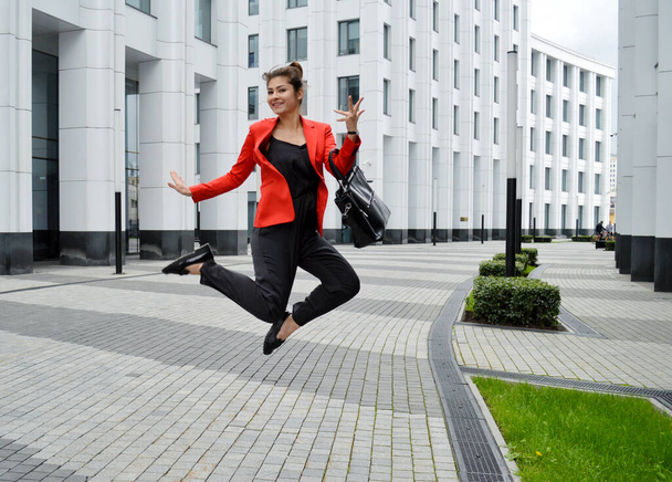 Beautiful business woman on the background of the modern office. A girl in a black overalls and a red jacket is jumping for joy near the business center - Fotografie, Obrázek
