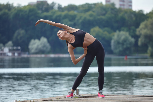 Attractive girl in sportswear does gymnastic exercises on a wooden pier in a city park. - Fotó, kép