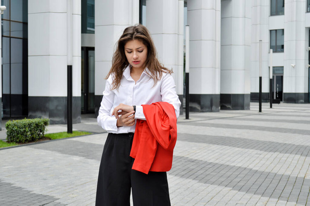 Beautiful business woman on the background of the modern office. girl with dark hair in a white shirt and black trousers with a red jacket in his hands looking at the clock near the business center - Foto, Imagen