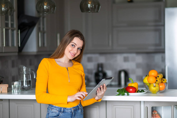 une fille dans un pull orange tient une tablette à la recherche d'une recette pour cuisiner des aliments sains - Photo, image