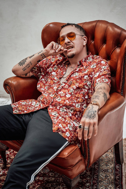 Очень выразительный и стильный парень в гавайской рубашке и спортивных штанах сидит на ретро-кресле, выглядит причудливо и безумно - Фото, изображение