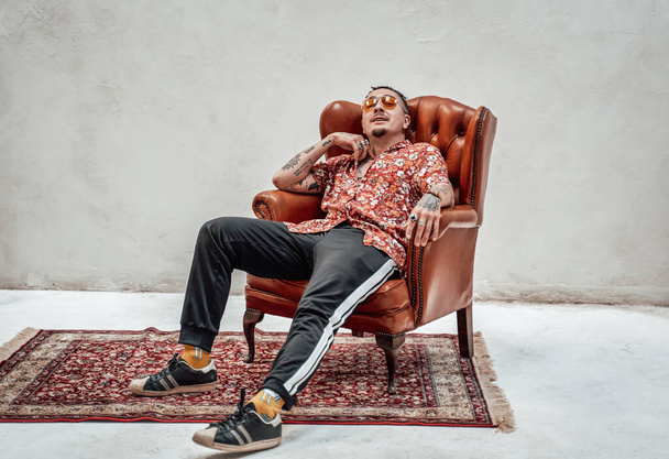 Very expressive and stylish guy in hawaiian shirt and sports pants sits on retro armchair, looks freakly and crazy - Photo, Image