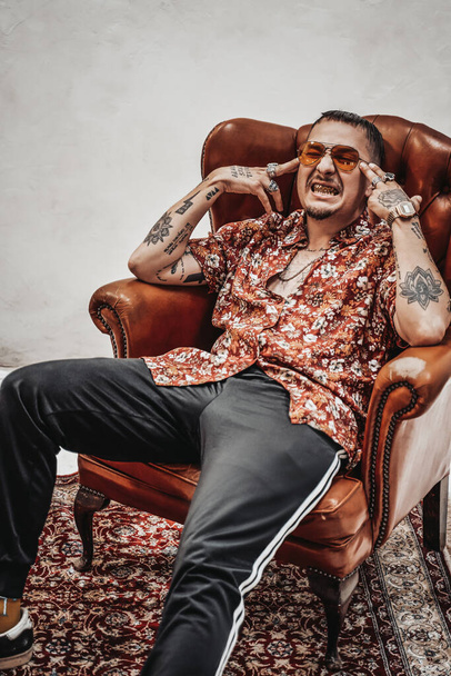 Expressieve en stijlvolle man in Hawaïaans shirt en zonnebril ontspannen op een luxe retro bank ziet eruit als freakly gangster - Foto, afbeelding