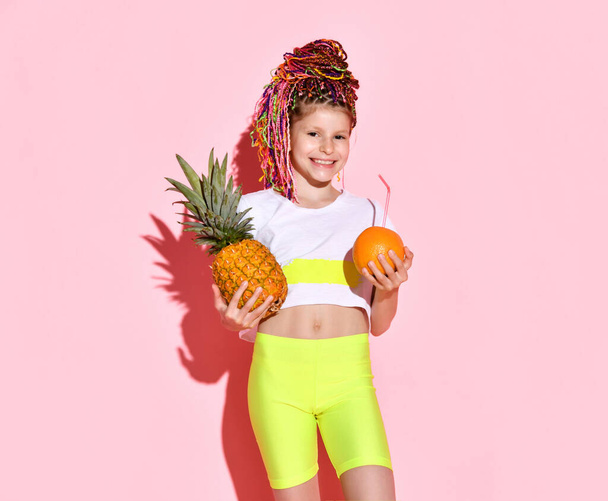 cute little girl with multi-colored pigtails on her head smiles happily, holding a pineapple and an orange in her hands. Cropped portrait isolated on pink, copy space. Childhood, emotions, summer. - 写真・画像