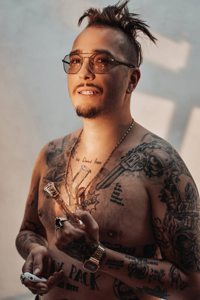Hombre sin camisa con el cuerpo tatuado y corte de pelo raro sostiene la pipa con cannabis - Foto, Imagen