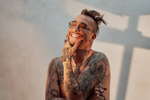 Retrato de un hombre desnudo y elegante en gafas de sol con cuerpo tatuado y accesorios, sonriendo con los ojos cerrados - Foto, Imagen