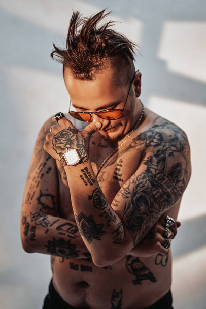 Muotokuva paidattomasta miehestä, jolla on ilmeikäs hiustenleikkaus ja tatuoitu runko aurinkolaseissa - Valokuva, kuva