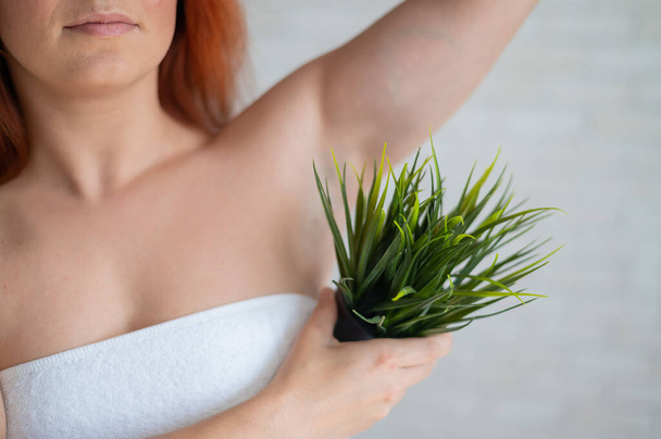 Close-up. Cropped. A woman in a white towel holds an armpit in a pot with a plant simulating unwanted vegetation on her body. Depilation. Hair removal in the armpit area. Smooth skin after epilation. - Foto, afbeelding