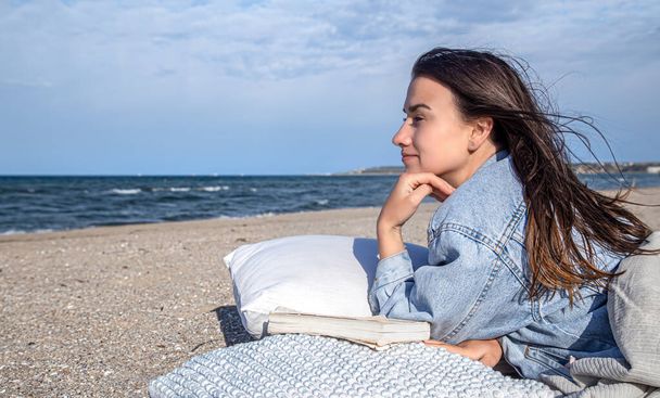 Relaks koncepcja, kobieta na plaży w wietrznej pogodzie spoczywa na poduszce z książką. - Zdjęcie, obraz