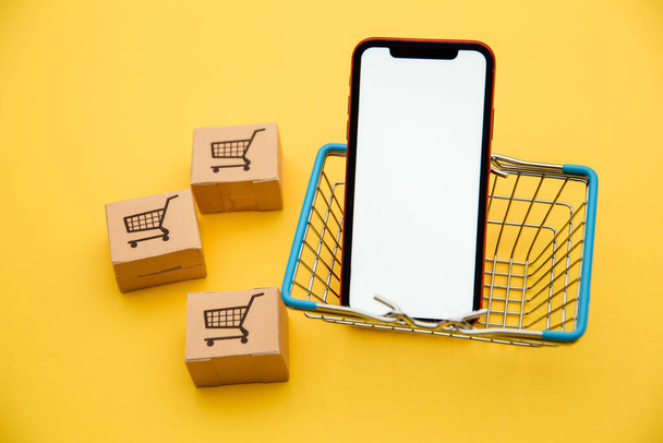 インターネットの概念を介してオンラインショッピングやeコマース:黄色の背景にバスケットに箱やスマートフォン - 写真・画像