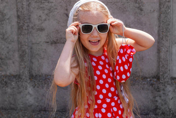 Krásná šťastná dívka v červené puntíkové šaty pózovat a usmívat se na betonové zdi pozadí. Roztomilé radostné dítě s dlouhými blond vlasy ve slunečních brýlích a bílý klobouk na šedém pozadí venku. Malý model - Fotografie, Obrázek