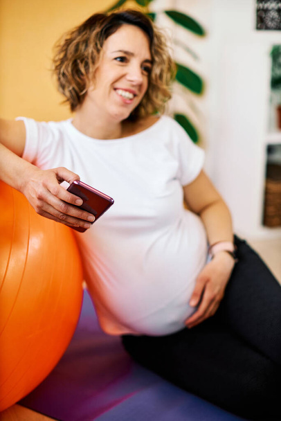 Middle aged fit pregnant sportswoman sitting on the floor at home, leaning on the pilates ball and using smart phone. - 写真・画像