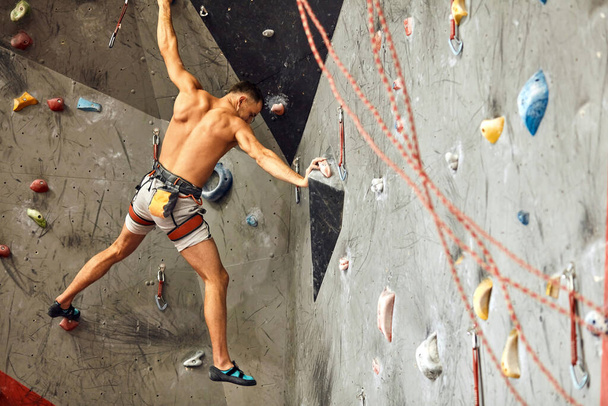 Alpinista masculino exercitando-se no ginásio, subindo na parede artificial com pedras
. - Foto, Imagem