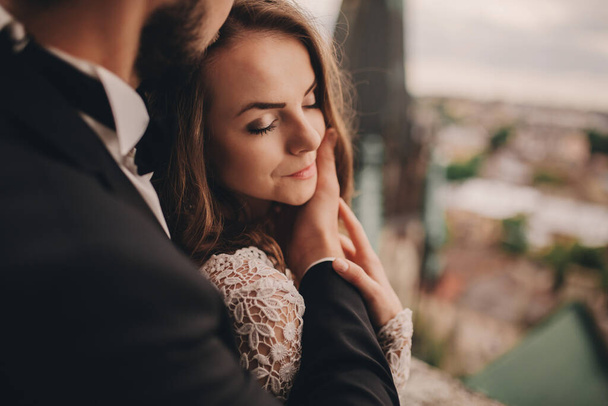 portrait of Happy newlywed. beautiful bride and stylish groom are hugging on the balcony of old gothic cathedral with panoramic city views. - 写真・画像