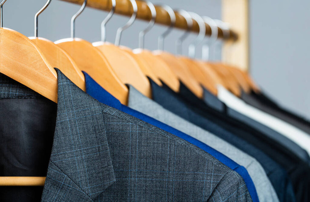 Suits for men hanging on the rack. Mens suits in different colors hanging on hanger in a retail clothes store, close-up. Mens shirts, suit hanging on rack. Hangers with jackets on them in boutique - Fotó, kép