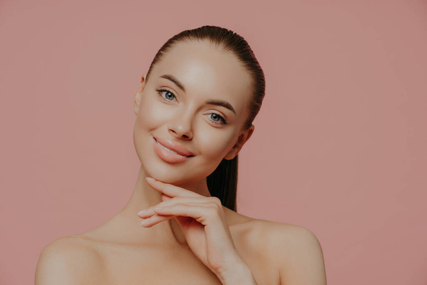 Charming beautiful woman with pony tail, touches perfect soft skin after cosmetology procedures, using beauty cream, stands with bare shouldes, poses against pink background. Natural as she is - 写真・画像