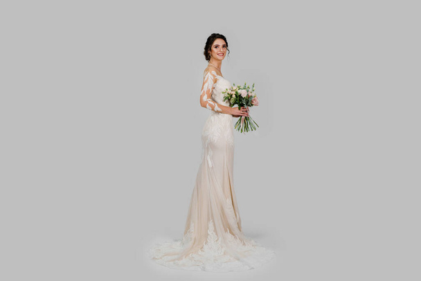Junge attraktive Braut mit Strauß im modernen Brautlook mit Kappenärmeln und einem Illusionsausschnitt runden die Säulensilhouette und die ausladende Schleppe ab. Braut Mädchen im Studio auf weißem Hintergrund - Foto, Bild