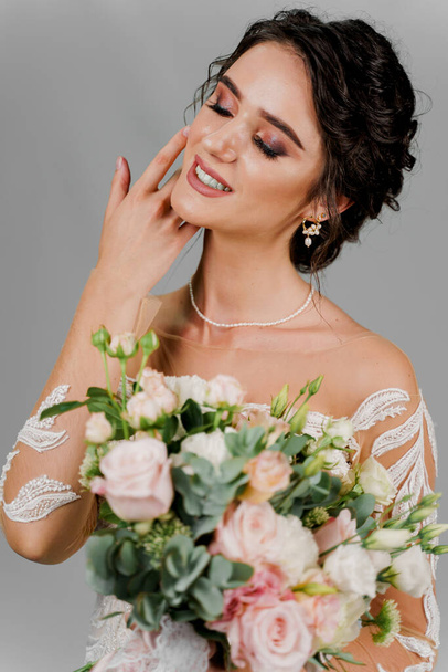 Bride with wedding bouquet smiles and touches her face and hair. Attractive girl portrait for social networks. Girl in wedding gown on blank background. Dreamily bride woman - Photo, Image