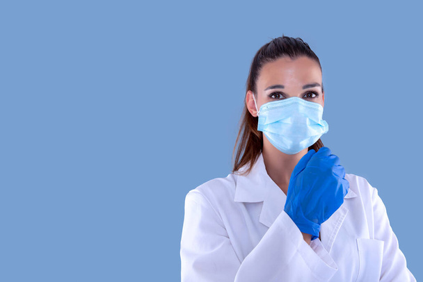 魅力的な女性医師とともにウイルス保護マスクとラテックス手袋をまっすぐ前と青の背景を見て. - 写真・画像