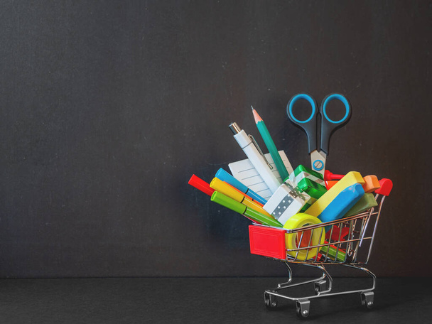 Colorful stationery and school bus model in shopping cart. Back to school concept. Vertical blackboard background with copy space on price tag. School shopping. - Foto, imagen