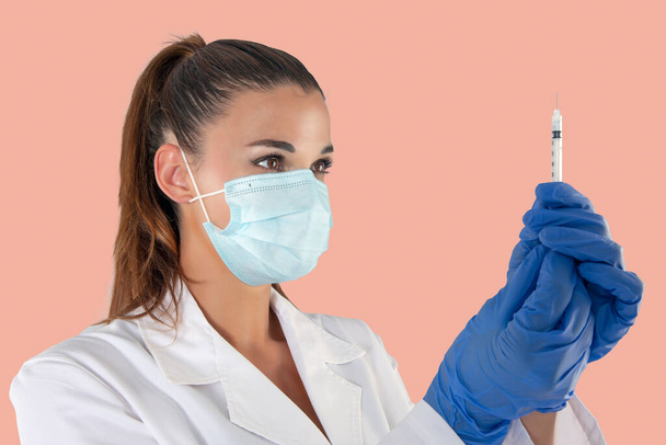 Ελκυστική γυναίκα γιατρός φορώντας μάσκα προστασίας από ιούς και γάντια λατέξ που φέρουν σύριγγα εμβολίου. - Φωτογραφία, εικόνα