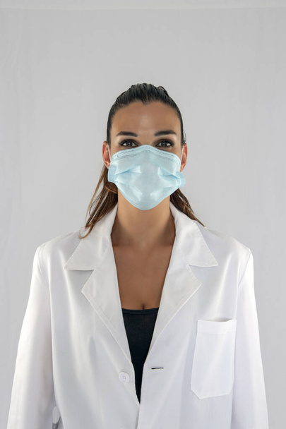 Atractivo médico femenino con máscara de protección contra virus mirando hacia adelante y fondo blanco. - Foto, imagen