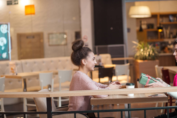 Happy woman congratulates her girlfriend with present box while sitting at cafe. - Photo, image