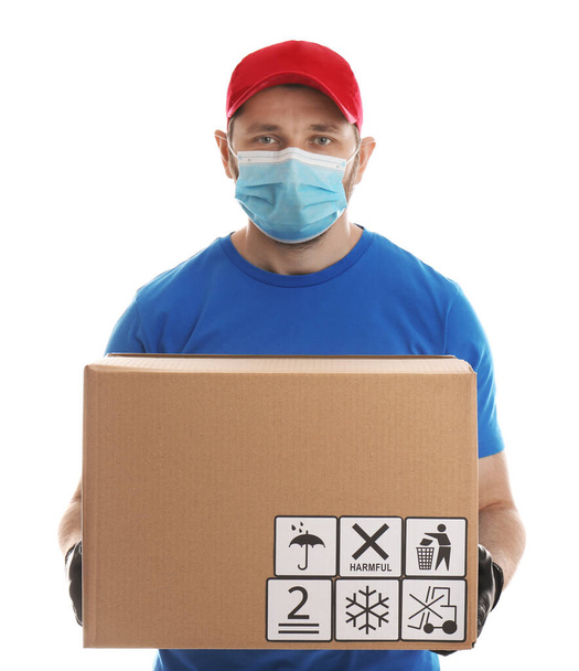 Courier in mask holding cardboard box with different packaging symbols on white background. Parcel delivery - Photo, Image