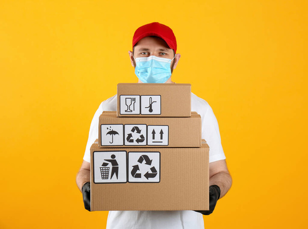 Courier in mask holding cardboard boxes with different packaging symbols on yellow background. Parcel delivery - Fotoğraf, Görsel
