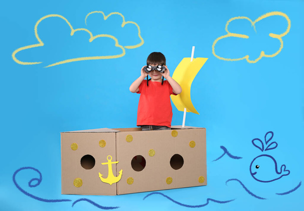 Cute little child playing in cardboard ship on light blue background with illustrations - Foto, afbeelding