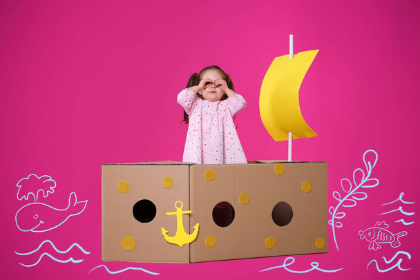Little child playing in cardboard ship on pink background with illustrations - Foto, imagen